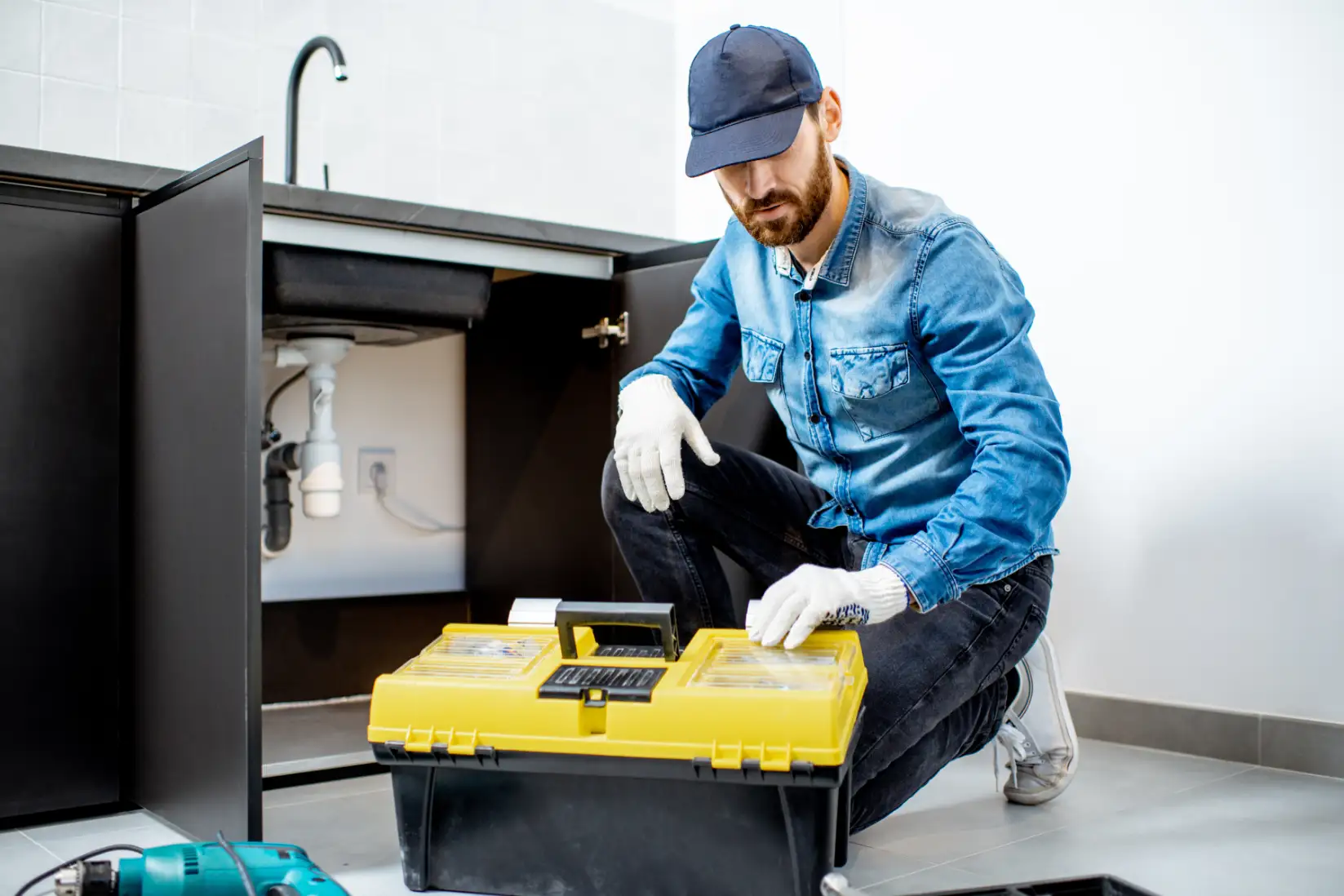 Plumber kneeling beside an open kitchen sink cabinet with a yellow toolbox