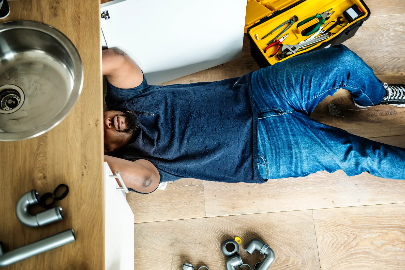 Plumber working beneath a kitchen sink with tools and pipe fittings nearby